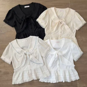 Flat lay overview of assorted sweet lolita short-sleeve sailor blouses in black, cream, and white with blue collar trim on wooden desktop, showcasing sailor collar with detachable bow tie, ruffled hem, and lace trim details