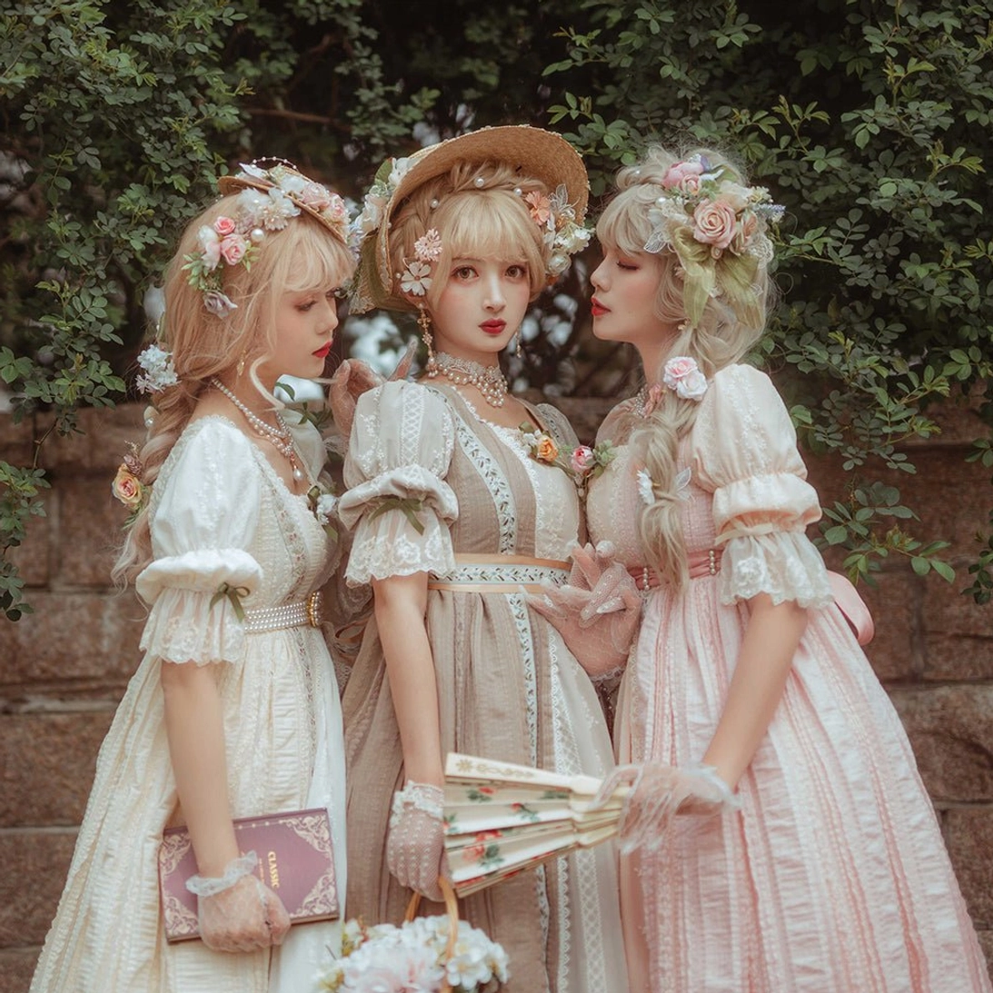 Three girls wearing classic empire waist lolita dresses with matching handmade ribbon belts and pearl embellishments in outdoor garden stone wall setting