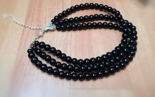 Three-strand black bead choker necklace on wooden table surface, showing opaque round black beads in three layered strands with silver lobster clasp and extension chain