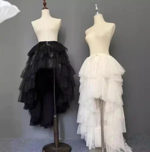 Front flat view of tiered high-low tulle half skirt in black and off-white color variants on wooden display stands showing ruffled layered tulle design and full silhouette
