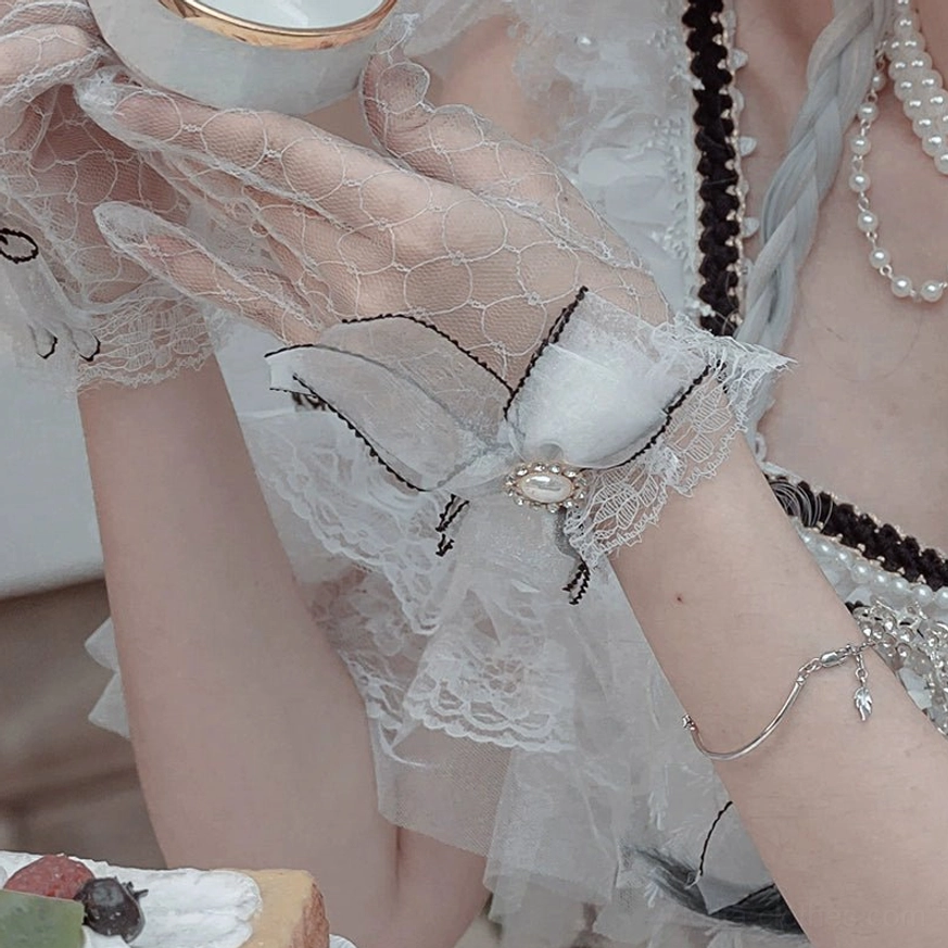 Transparent mesh wedding Lolita gloves with black ruffled edges, ribbon bow wrist detailing, satin bow and gem brooch accents worn by model holding porcelain teacup, showing delicate lace cuff and sheer mesh construction in top-down hand perspective