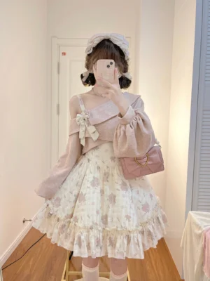 Twilight Rose sweet lolita two-piece set featuring pink floral print high-low JSK dress with adjustable shoulder straps and dusty pink off-shoulder sweater with satin bow accent, modeled with twin buns hairstyle and matching pink heart-shaped handbag