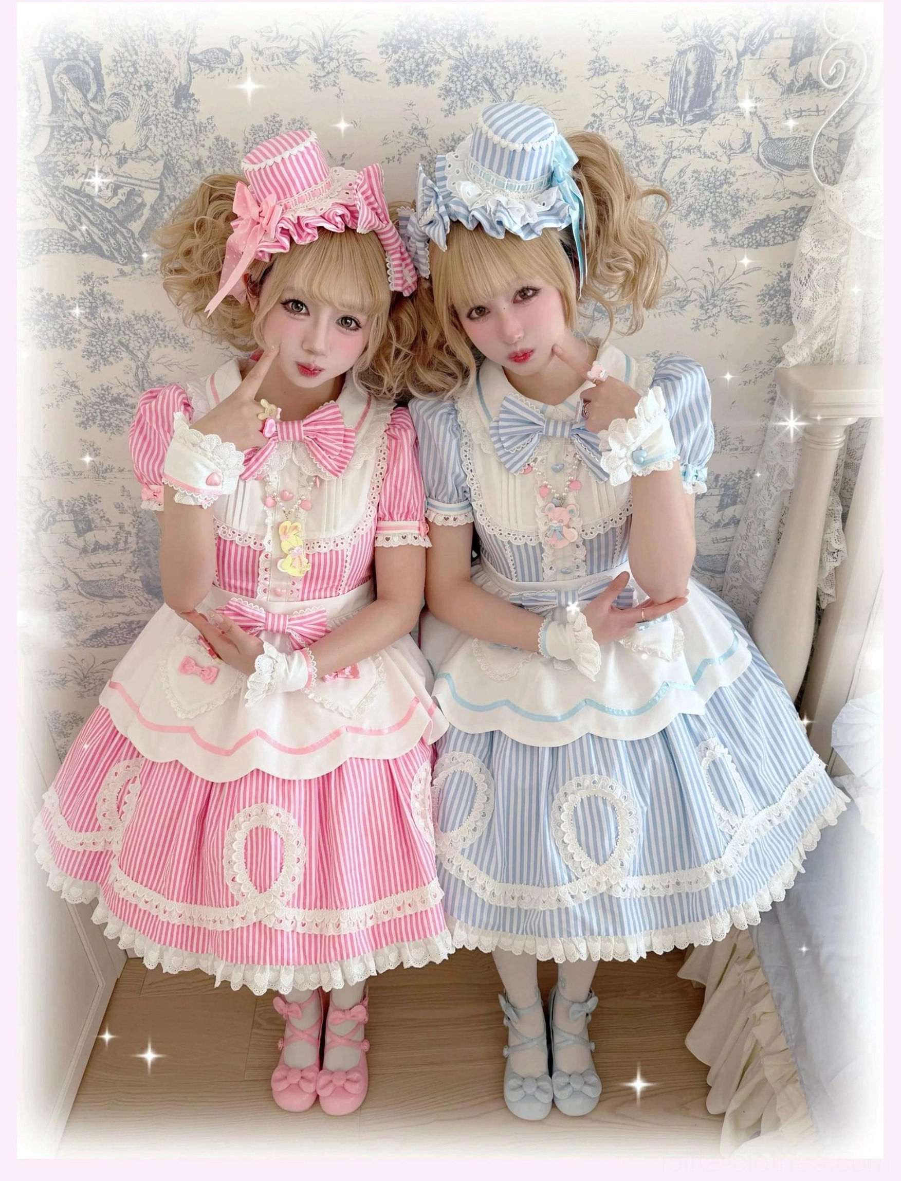 Two female models in pink and blue striped lolita OP dresses with sparkly star effects wearing matching bonnets and heart-shaped button cuffs