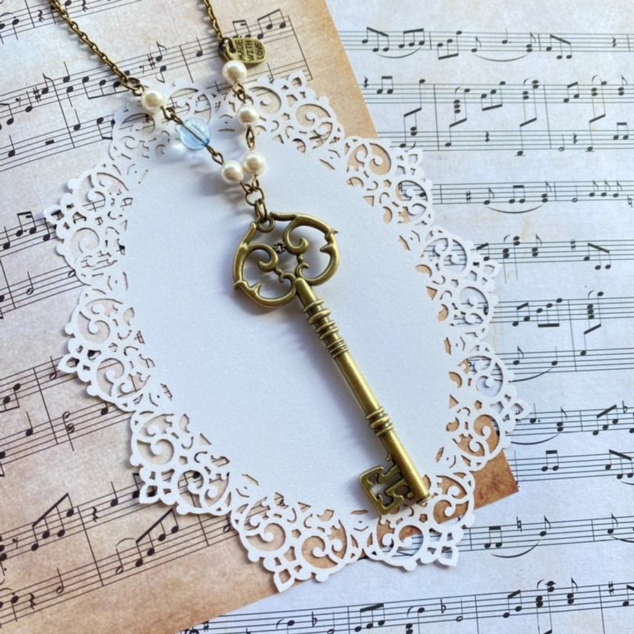 Vintage bronze key-shaped Lolita necklace sweater chain with artificial pearls and light blue glass beads displayed on sheet music and white lace doily flat lay background