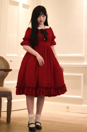 Vintage classic lolita OP dress burgundy red color white doll collar with black bow tie ruffled midi skirt full body front standing view on model with long black hair minimalist indoor wooden chair background