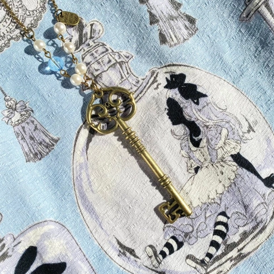 Vintage key long necklace with large bronze key pendant paired with pearl and blue crystal beads on Alice in Wonderland themed printed fabric background