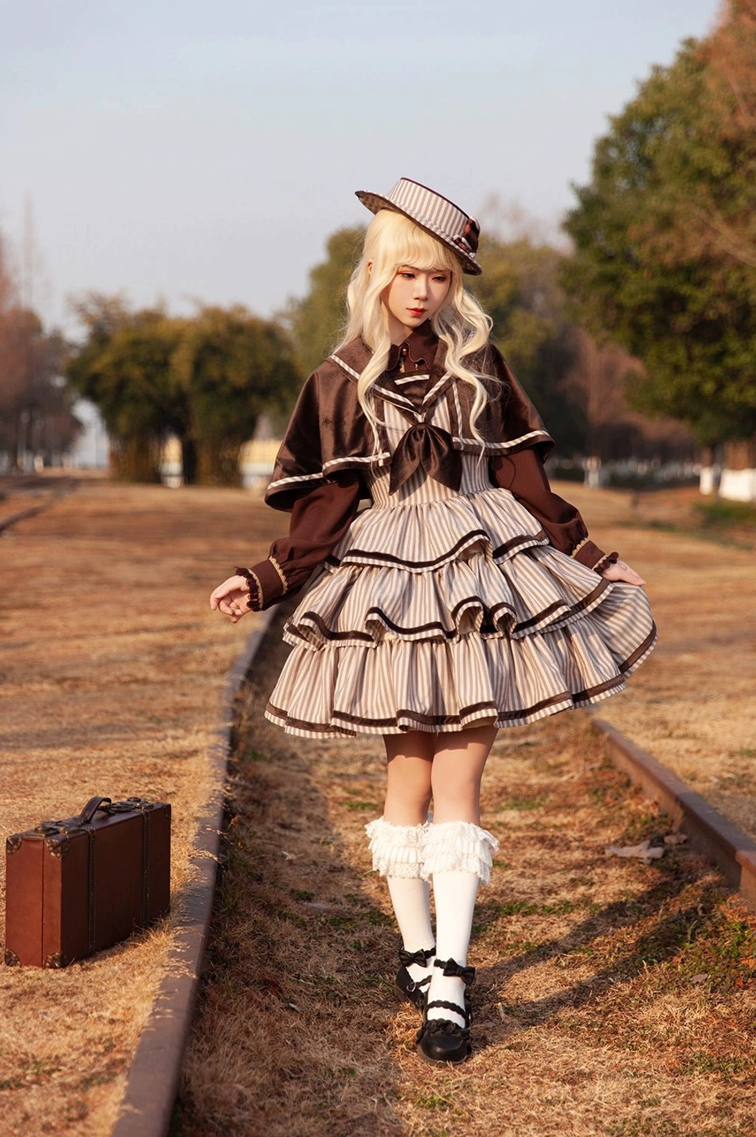 Full-body front walking shot of blonde model in vintage striped peppy sailor Lolita JSK with brown cape walking on railway track, featuring vintage leather suitcase accessory, ruffled socks, and loafers in retro outdoor railway setting