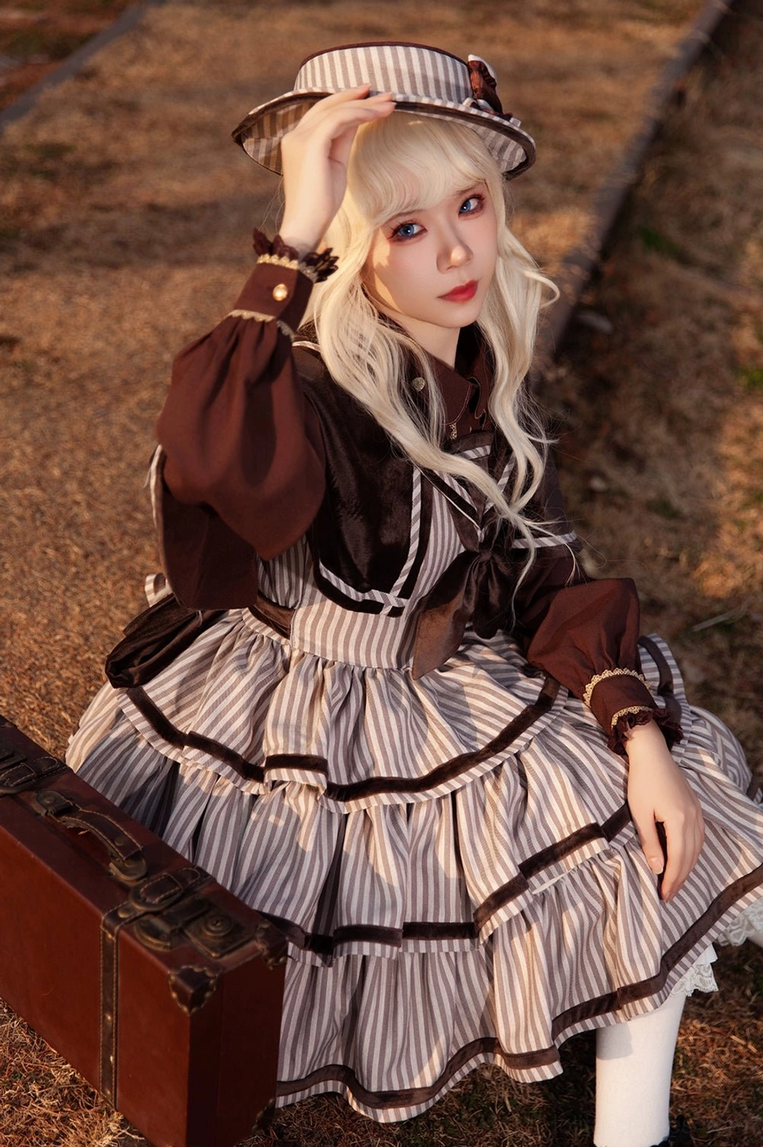 Close-up front seated shot of blonde Lolita model wearing vintage striped peppy sailor JSK with dark brown long-sleeve inner top, matching sailor hat, and elegant retro styling details at railway platform side