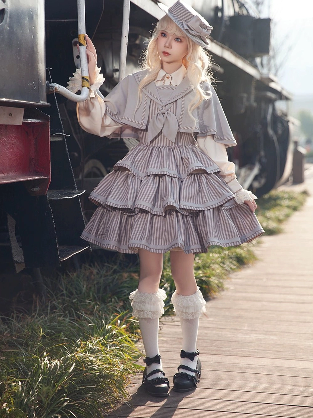 Full-body model wearing grey-brown striped vintage peppy sailor Lolita JSK coordinate beside vintage train wooden walkway, styled with matching mini sailor hat, ruffled white socks, and leather loafers for authentic period aesthetic