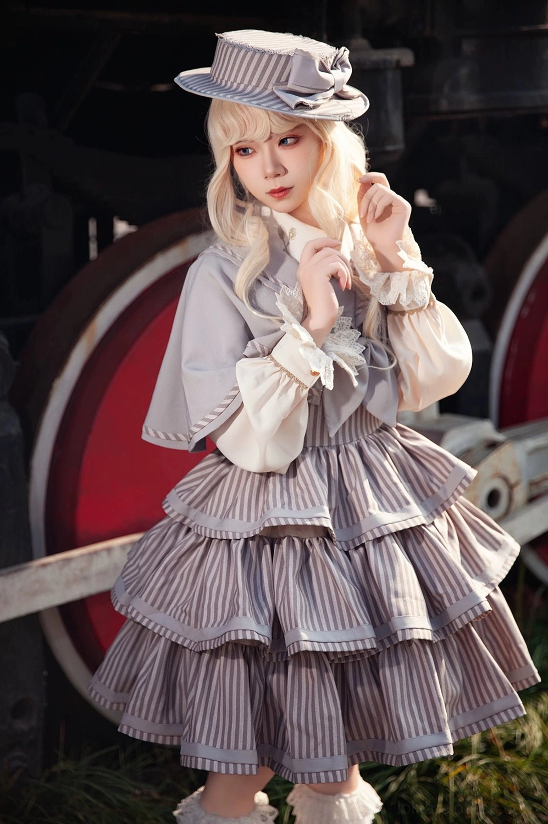 Half-body shot of blonde Lolita model wearing grey-brown striped JSK with puffed ruffled sleeves and decorated sailor hat beside vintage train wheel, showcasing full layered cake skirt design and structured silhouette