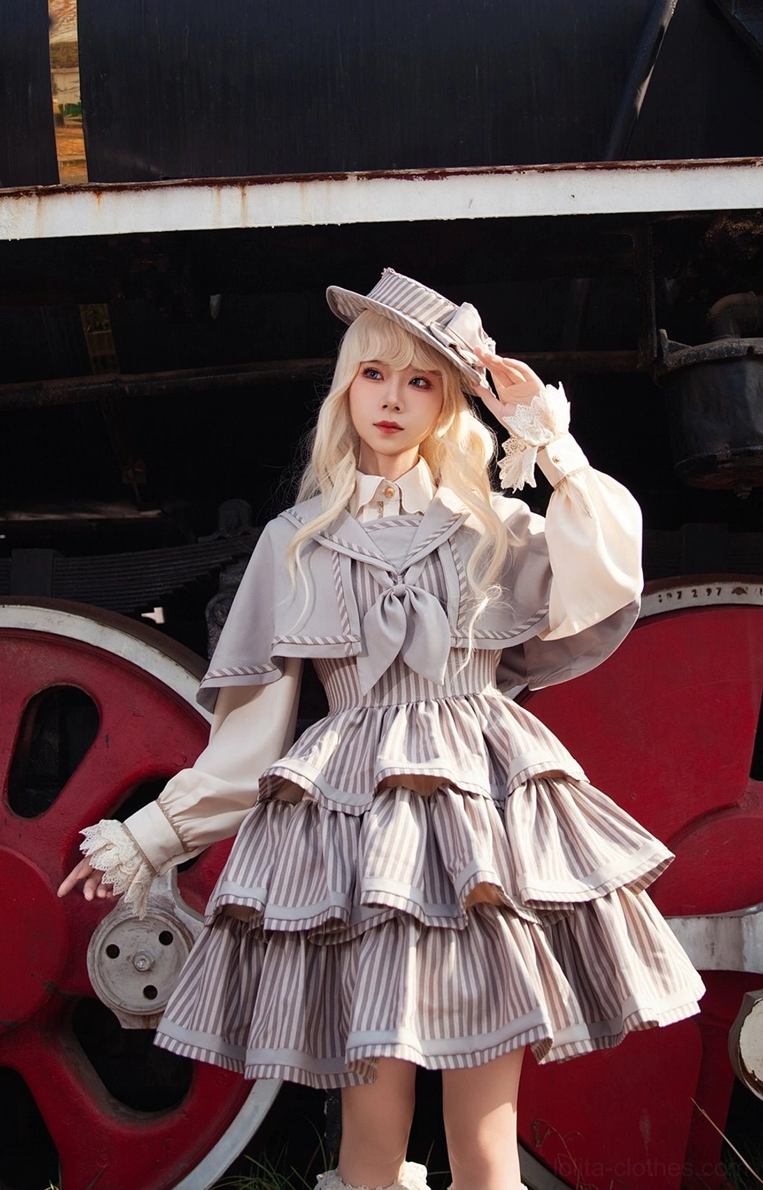 Full-body side view of model in vintage striped grey-brown peppy sailor Lolita JSK leaning against vintage train wheel, showcasing decorated sailor hat, elegant ruffled cuffs, and layered cake skirt silhouette with posed styling