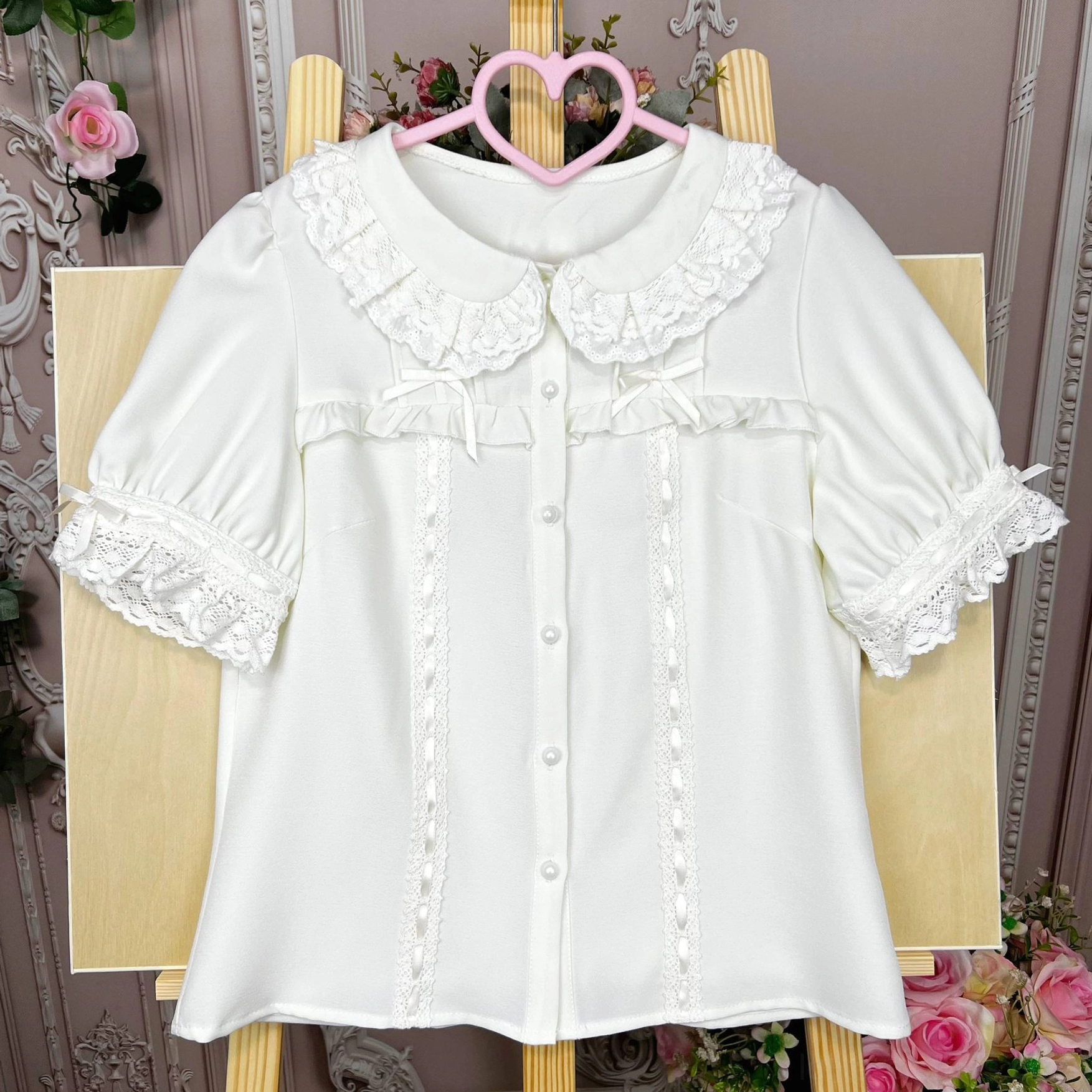Lolita blouse with ruffled doll collar and chest ribbon bows displayed on heart hanger against retro rose European backdrop in side front hanging view