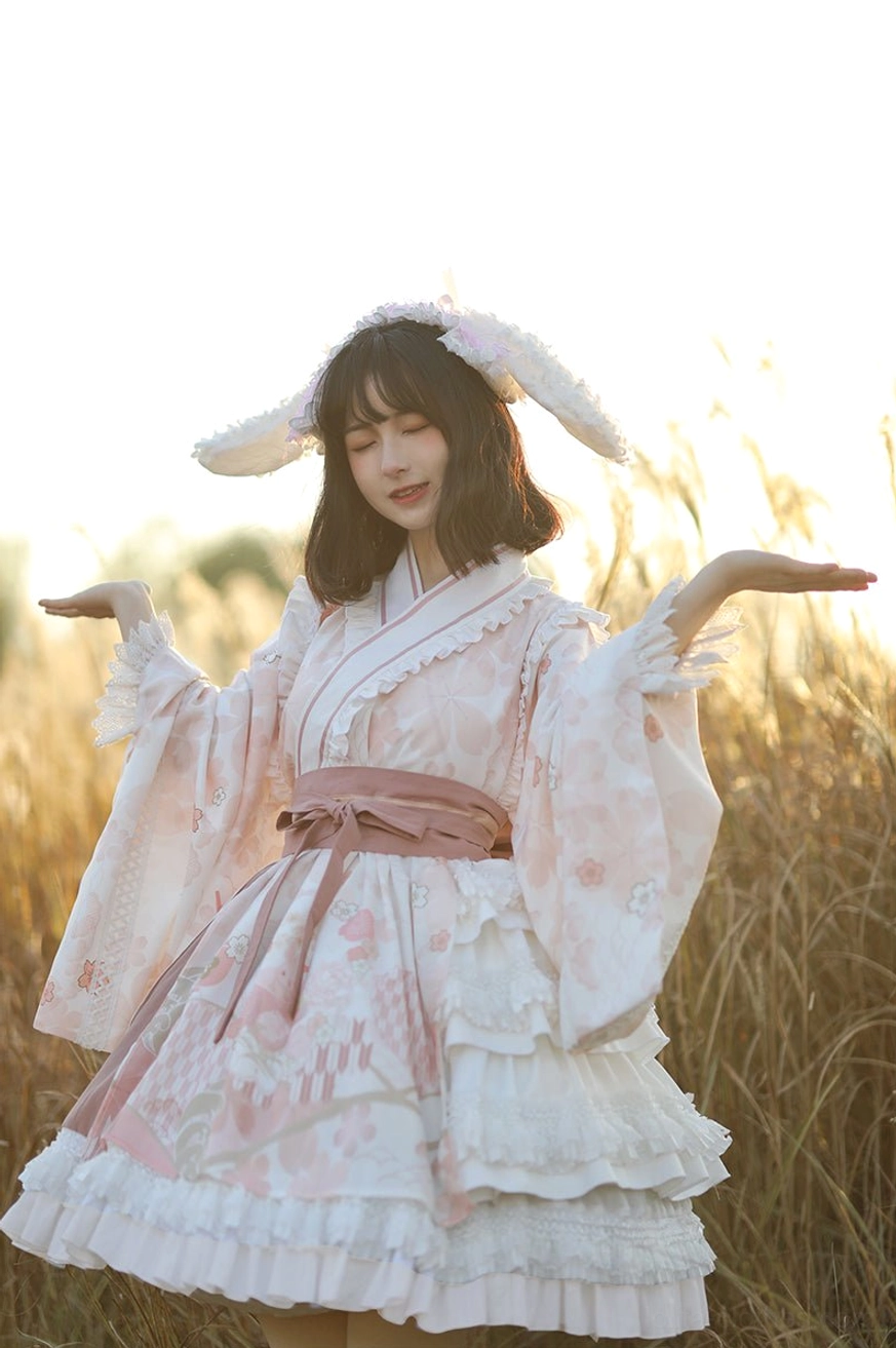 Cherry blossom print Wa Lolita OP dress with pink waist sash and rabbit ear hair clips, front eye-level shot with model arms raised in sunset reed field showing layered ruffled skirt details