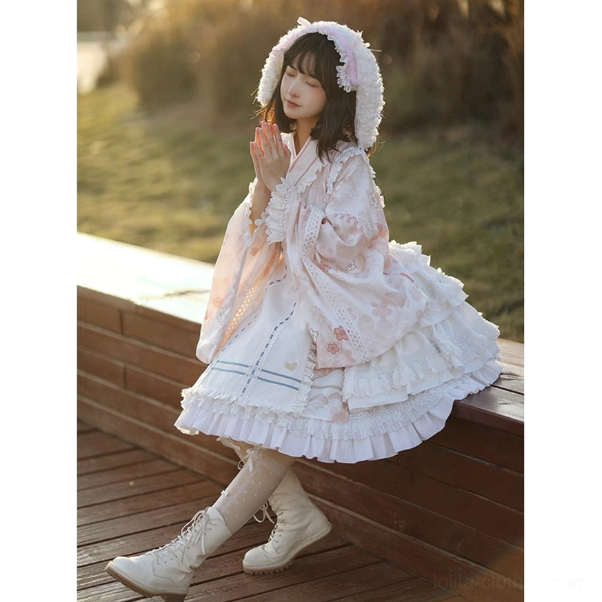 Kimono-style Lolita OP dress with hooded design and pink floral print, three-quarter side view of model sitting on wooden platform at sunset with tiered ruffled hem, white lace trims, and furry hood