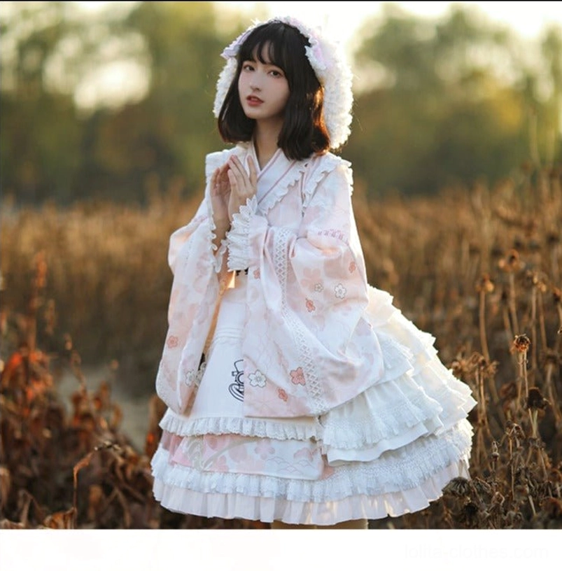 Short kimono-style Lolita coat layered over OP dress with pink floral print and matching furry hood, three-quarter front view in dry grass sunset scene with ruffled skirt hem and white lace trim