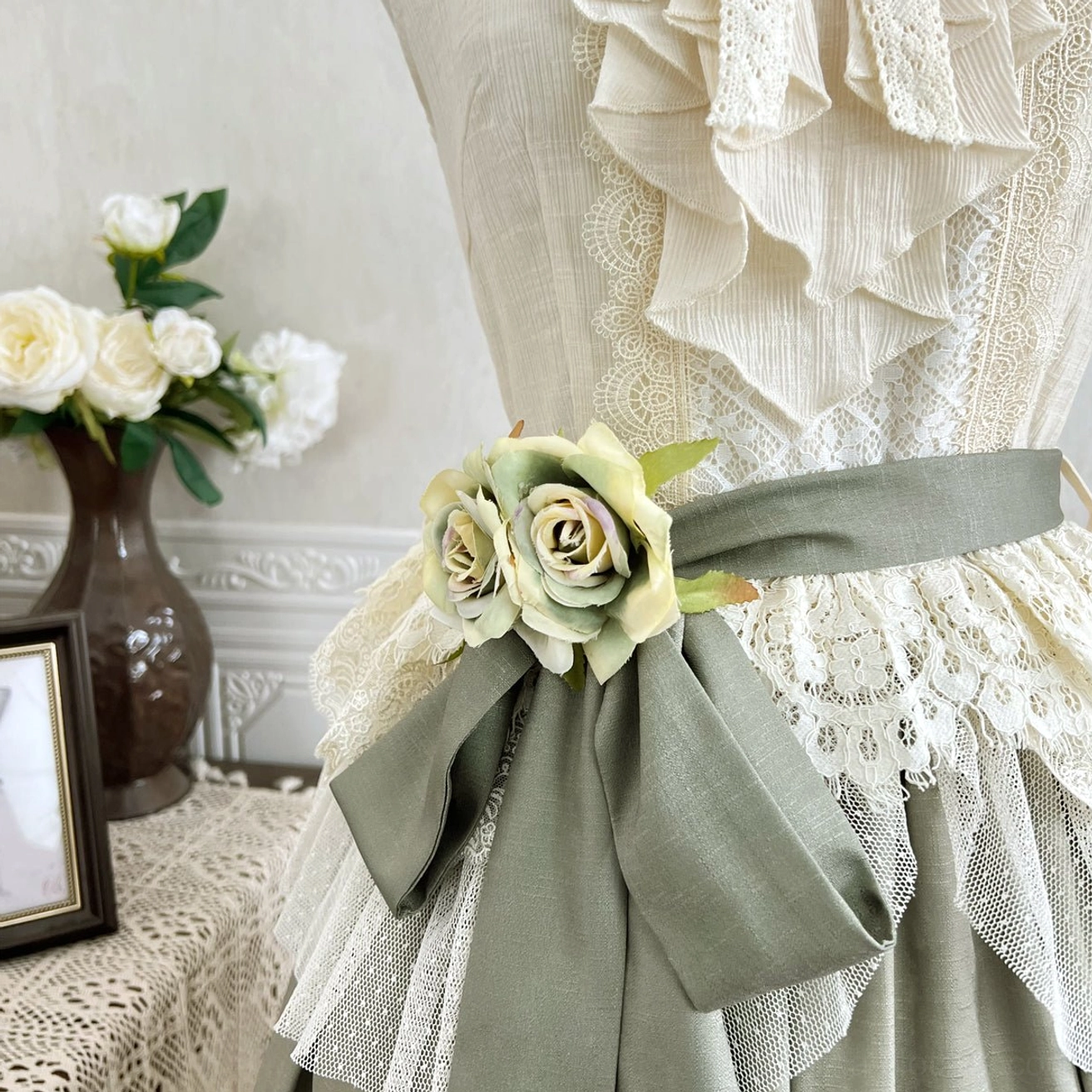 Side view close-up of large sage green fabric waist bow with dual rose flower decorative accents and layered ruffled blouse trim underneath
