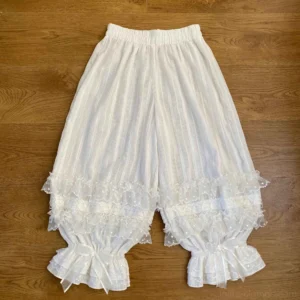 White cotton Lolita bloomers with vertical striped fabric base and multi-layered polka dot tulle ruffled cuffs featuring satin ribbon bows, displayed in full flat lay view on wooden surface