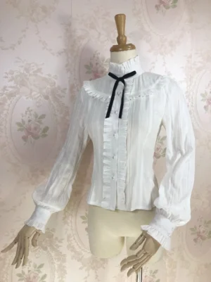White ruffled stand collar lolita blouse with black velvet bow on dress mannequin, front three-quarter view showing lantern sleeves and pearl button details