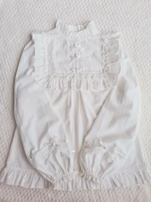 White cotton lolita long-sleeve blouse with ruffled collar, square lace yoke, pearl buttons, and satin bows displayed on flat lay - sweet lolita fashion piece