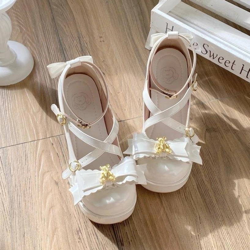 White sweet lolita platform heels flat lay side shot on wooden floor, decorative sweet home sign in background, patent leather finish, crisscross ankle straps, ruffled bow with gold bear charm, transparent block heel