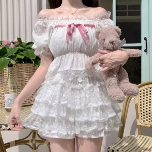 White multi-layered floral Lolita bloomer ruffled shorts with pink chest bow and off-shoulder puff sleeves worn by model in outdoor French patio setting holding teddy bear