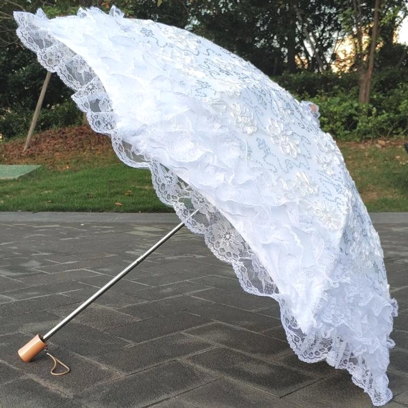 White lace Lolita sun parasol with tiered ruffled lace trim and wrist strap handle photographed outdoors in garden patio setting with green plants background