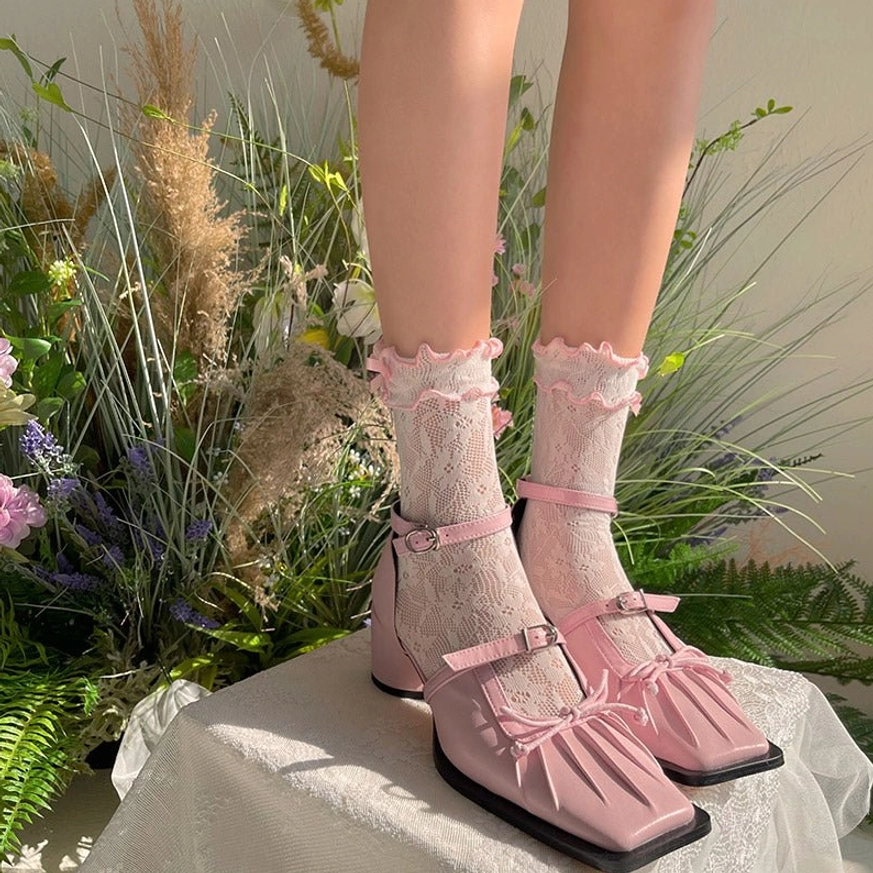Delicate white sheer lace Lolita ankle socks with pink ribbon bow paired with pink square-toe Mary Jane shoes displayed on model's feet with floral greenery background