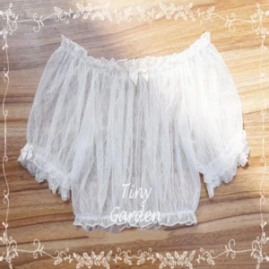 White off-shoulder cropped lolita blouse with sheer rhombus mesh fabric and ruffled lace hem, featuring tiny bow detail at boat neckline, flat lay top-down view on wooden surface
