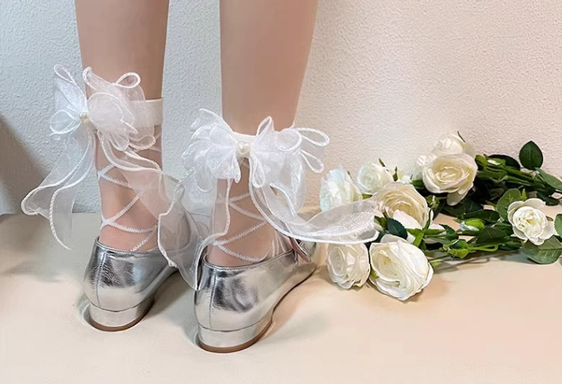 Front lower leg and foot view of pair of white lolita ankle socks with delicate organza bows and sheer lattice crisscross straps worn with silver platform shoes on white rose table decor