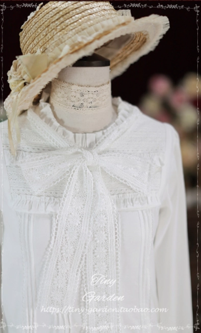Front close-up of pure white Lolita long-sleeve blouse showing large lace bow tie, full lace front decorative panel, and ruffled stand collar detail against retro floral studio backdrop