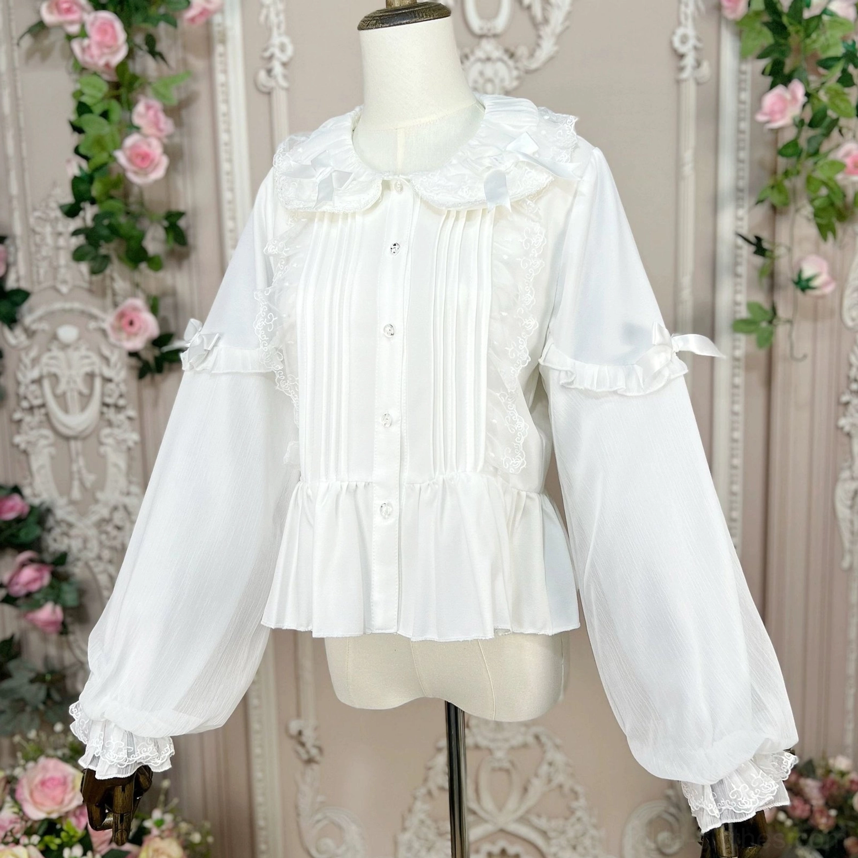 Front view of white Lolita blouse on dress form showing Peter Pan collar with ruffled details, front lace pintuck trim, decorative sleeve bows and ruffled hem accents - vintage floral wall background