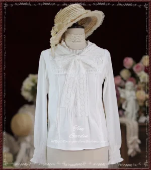 Pure white Lolita long-sleeve blouse featuring large lace bow tie at neck, ruffled stand collar, front pleated panels, and chiffon sleeves on dress form with retro flower arrangement studio backdrop