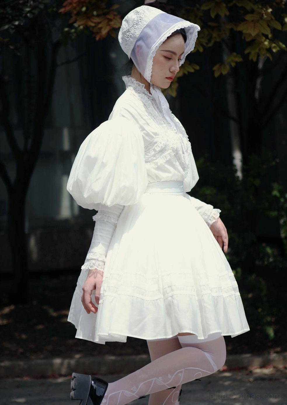 Side three-quarter view of female model wearing white Victorian Lolita blouse and mini skirt set with matching white bonnet, layered ruffled cuffs, slim long under-sleeves, and high-waisted A-line skirt outdoors