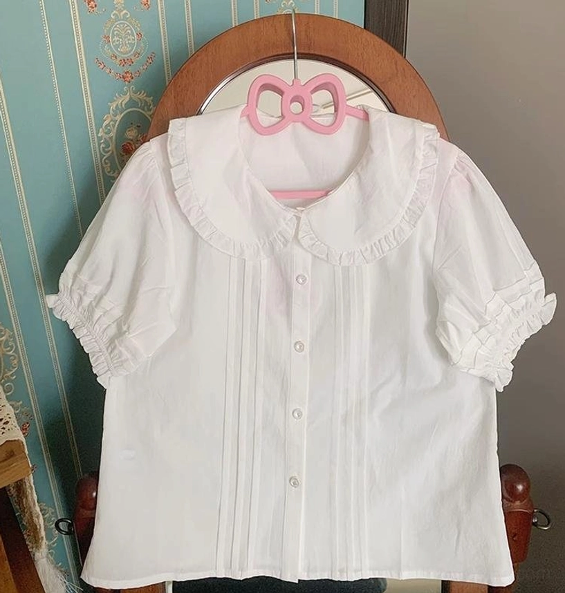 White short sleeve lolita blouse with classic peter pan ruffled collar, puff sleeves and front pleats displayed on pink bow hanger against teal vintage wall background