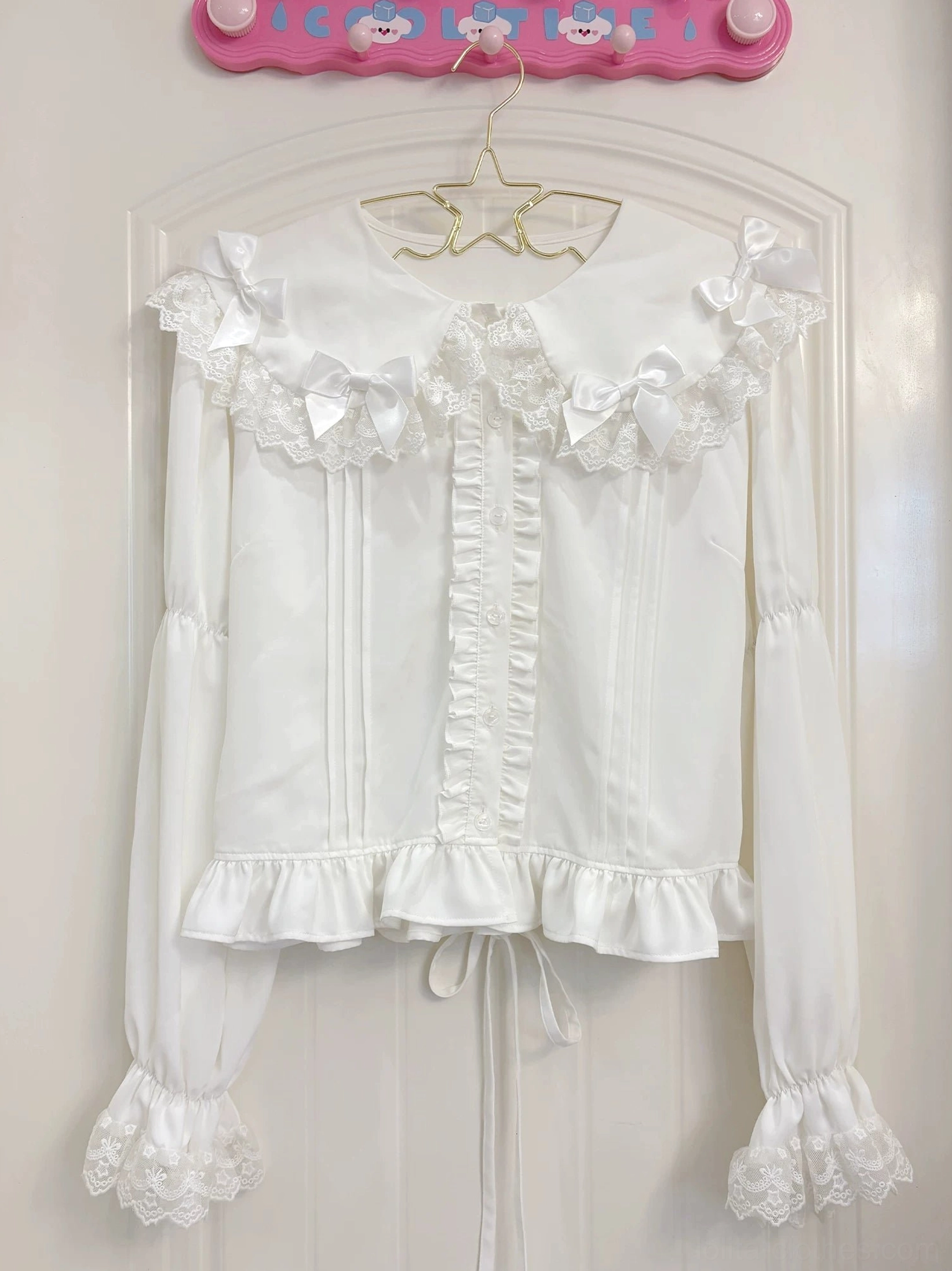 Front view flat lay product shot of white Lolita blouse featuring Peter Pan collar with lace and white bows, front ruched trim, and ruffled cuffs on star-shaped hanger