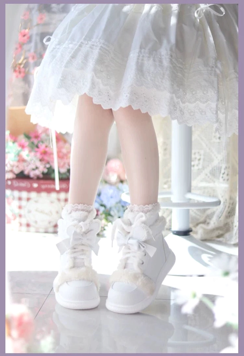 All-white lolita winter ankle boots with fluffy ankle trim and bow lace fasteners, low frontal view worn on model with floral studio backdrop