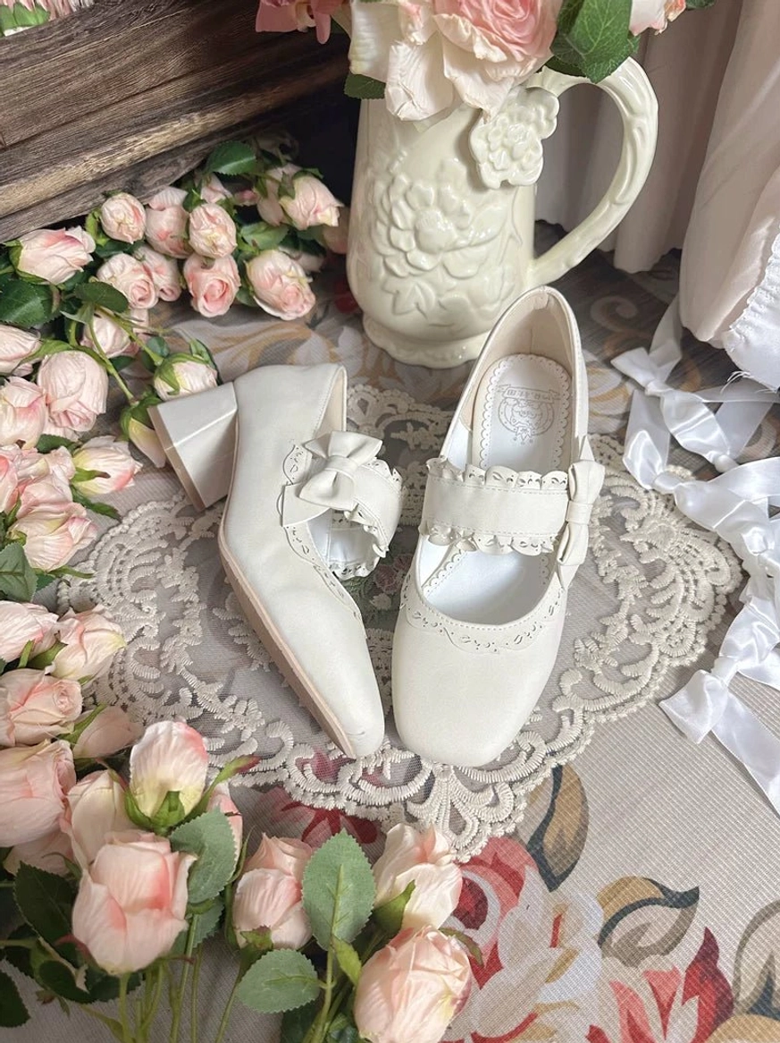 Pure white classic lolita chunky heel mary jane shoes with decorative bow ornament on ankle strap and embroidered vamp trim, flat lay styled with pink roses lace doily and vintage porcelain pitcher