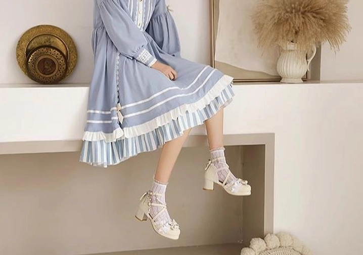 Side view of white Lolita heeled sandals with lace cross straps bow and pearl accents block heel worn by model sitting on shelf in minimalist interior setting with lace socks