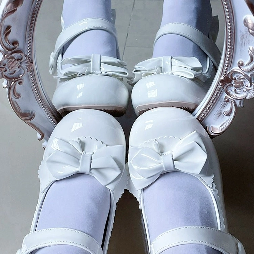 White Lolita Mary Jane patent leather shoes with large glossy toe-cap bow smooth patent surface heart-shaped strap buckle and round closed toe flat lay close-up beside vintage mirror