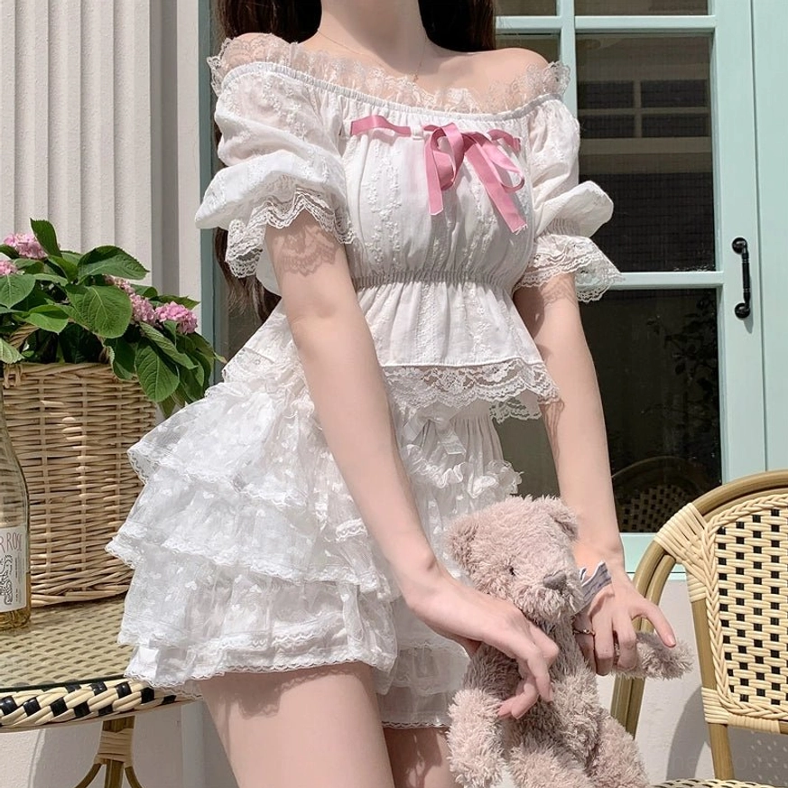 White Lolita ruffled two-piece set with shirred cropped top, layered tulle cake shorts and pink chest bow detail worn by standing model on outdoor French patio full waist-to-thigh view
