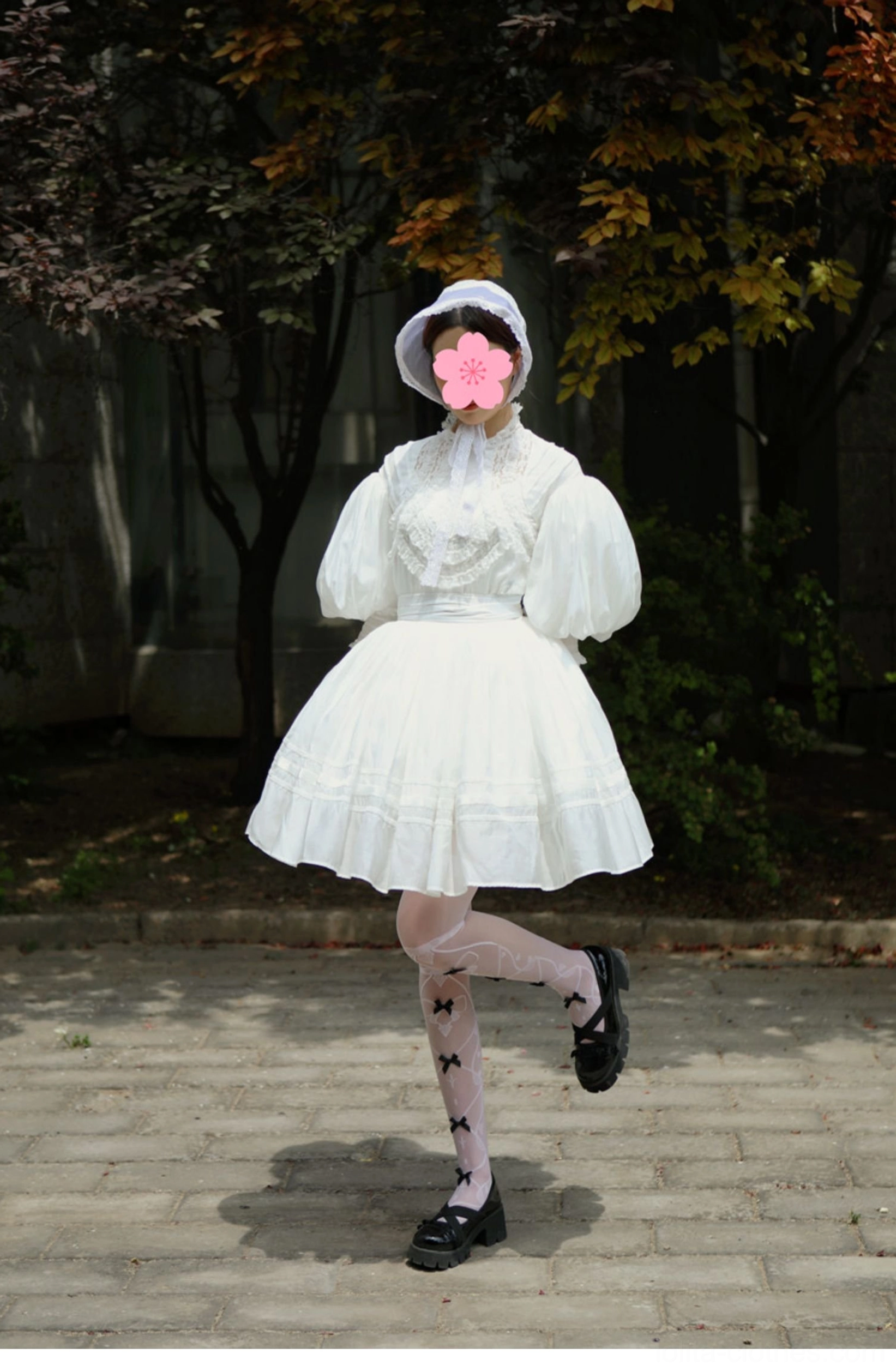 Front full view of model wearing short white Victorian Lolita set with white bonnet featuring flower face cover, short puff sleeves, high-waisted A-line mini skirt, and star-patterned sheer stockings outdoors