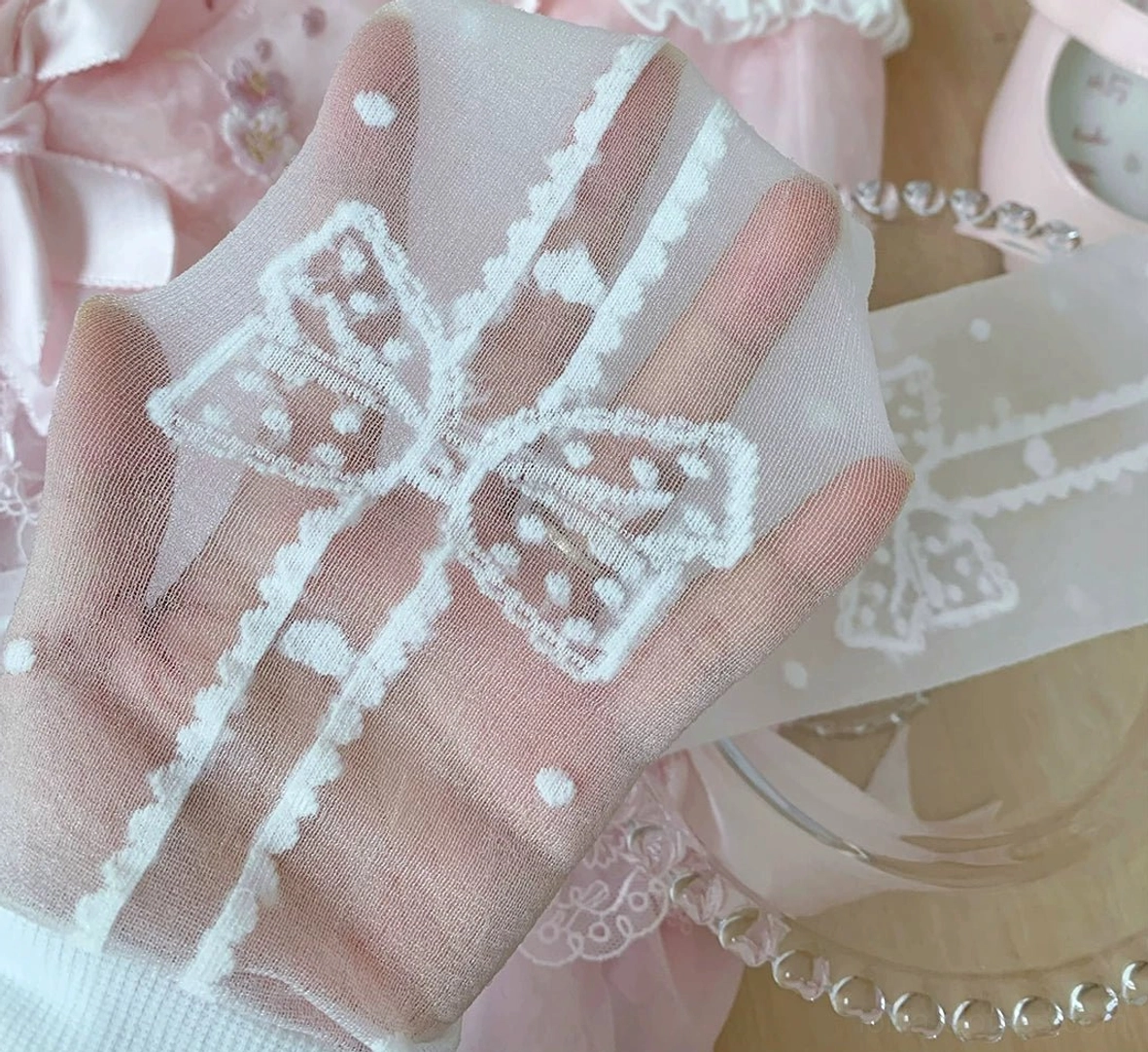 Close-up detailed handheld flat lay of white bow polka dot lolita socks showing delicate embroidered bow motifs sheer tulle material soft lace texture and intricate pattern details on pink lace background