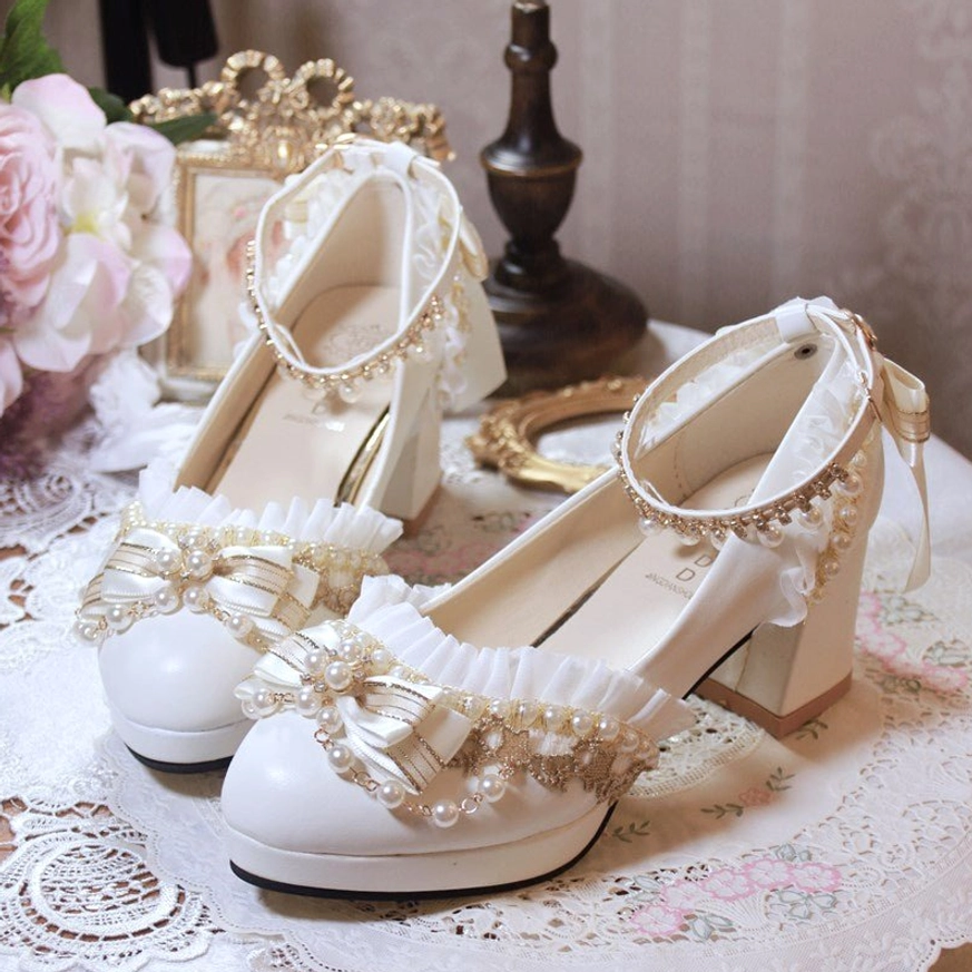 White handmade Lolita wedding heel shoes featuring pearl and gold beaded bows displayed on vintage lace tablecloth with pink roses and retro ornaments three-quarter front view