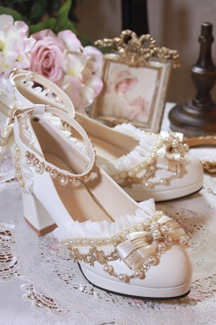 Two individual white handmade Lolita wedding heels high side angle view featuring exquisite pearl and gold beaded bow ruffled ankle strap clean white shoe body vintage setting