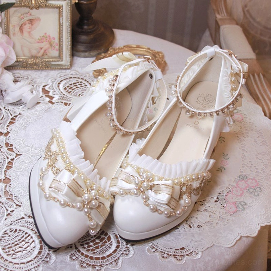 White handcrafted Lolita wedding heel shoes top-down three-quarter view on round vintage table showing ankle buckle strap ruffled shoe opening elegant bow with gold beaded details