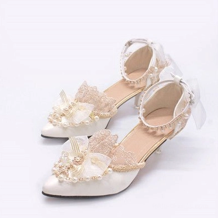 White pointed toe Lolita bridal wedding heels overhead flat lay, showcasing pristine white satin upper, white bow with pearl flower decor, intricate lace ruffles, pearl ankle strap, gold trim accents, and refined stiletto heel