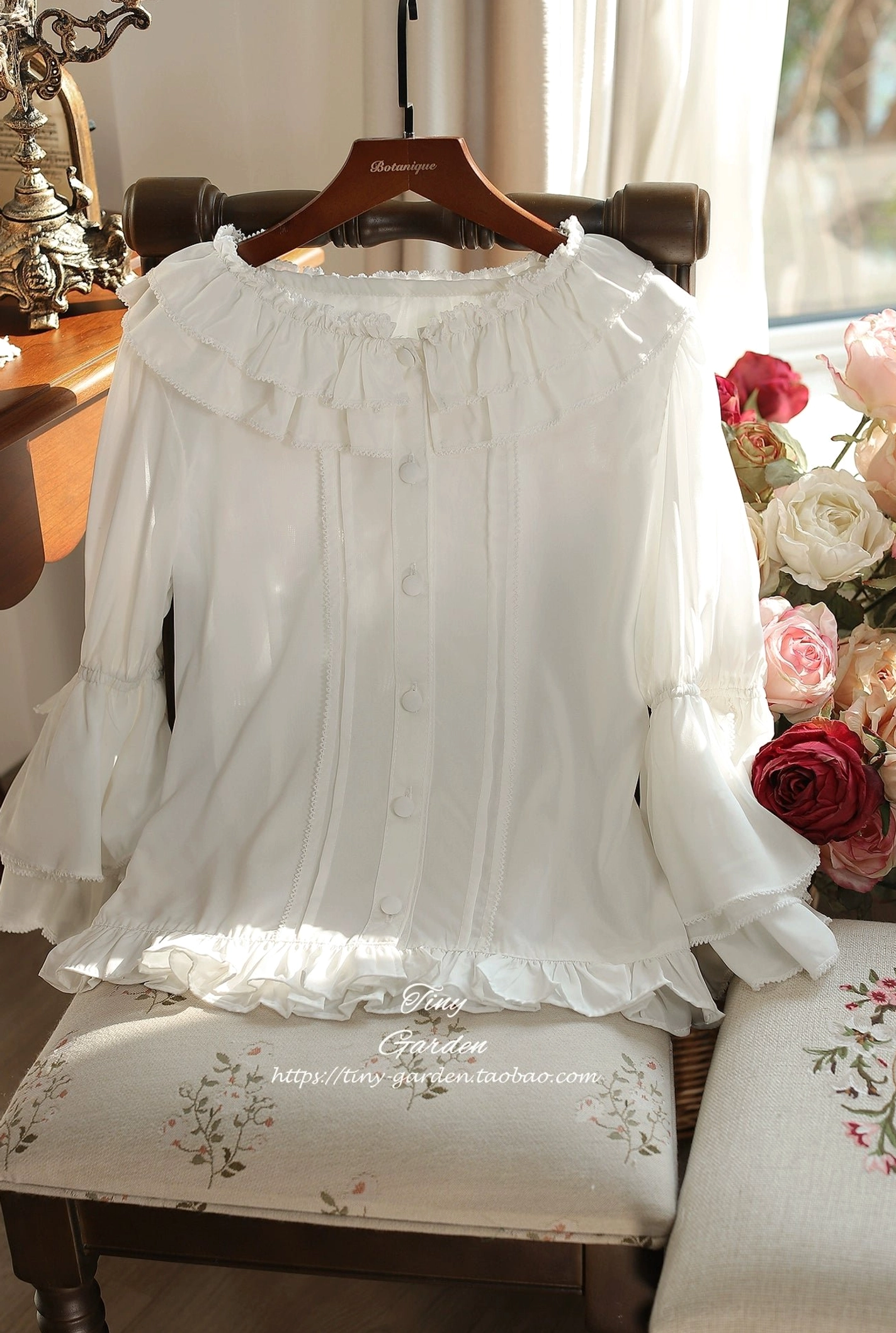 Minimalist white ruffled collar lolita blouse with simple layered bell sleeves, hanging on chair display with flower decoration background, round ruffled collar and front button placket