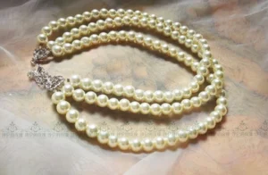 Close-up flat lay of three-strand white pearl choker on soft fabric, featuring cream-colored round pearls, silver lobster clasp, and adjustable extension chain detail