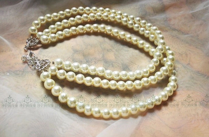 Close-up flat lay of three-strand white pearl choker on soft fabric, featuring cream-colored round pearls, silver lobster clasp, and adjustable extension chain detail