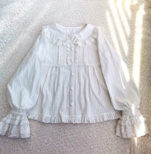White long-sleeve lolita cotton blouse with peter pan lace collar, tiered ruffle cuffs, and pleated front detailing, displayed on lace fabric with sunlight shadows, showcasing relaxed a-line silhouette and vertical striped cotton texture